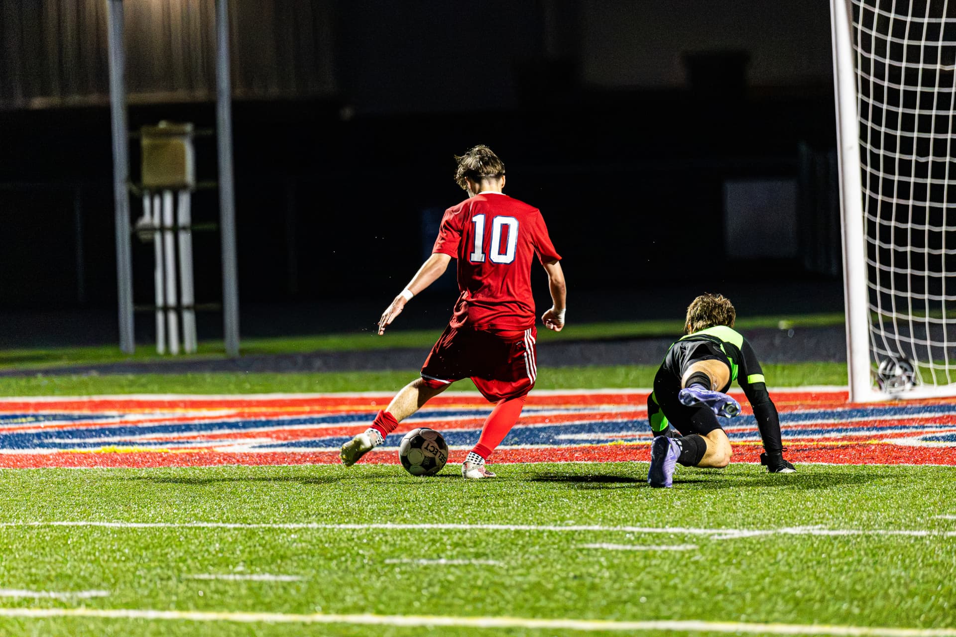 High school soccer striker in red scoring past the goalkeeper at night — soccer action photography by Striide